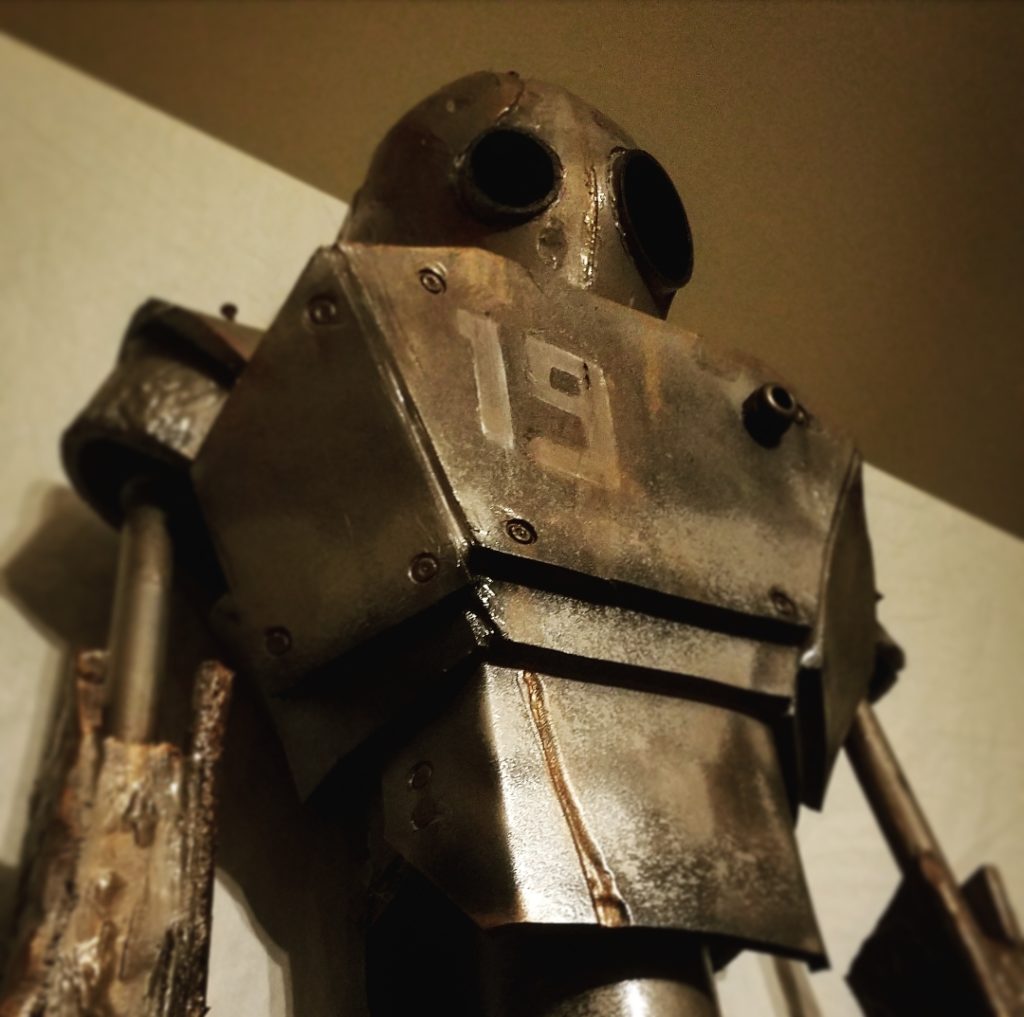 A robot stands 6ft tall with the number 19 displayed on its chest. There is rust coming from the edges of the plating. There are dents and scratches covering the robot’s head, arms, and chest. It seems to be looking at something in the distance with dark, black lenses–one of them larger than the other. The background is white, bringing a dynamic contrast to the dark bronze metallic glow of the robot’s plating.