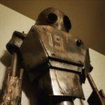 A robot stands 6ft tall with the number 19 displayed on its chest. There is rust coming from the edges of the plating. There are dents and scratches covering the robot’s head, arms, and chest. It seems to be looking at something in the distance with dark, black lenses–one of them larger than the other. The background is white, bringing a dynamic contrast to the dark bronze metallic glow of the robot’s plating.