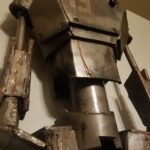 A robot stands 6ft tall with the number 19 displayed on its chest. There is rust coming from the edges of the plating. There are dents and scratches covering the robot’s head, arms, and chest. It seems to be looking at something in the distance with dark, black lenses–one of them larger than the other. The background is white, bringing a dynamic contrast to the dark bronze metallic glow of the robot’s plating.