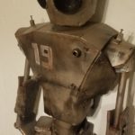 A robot stands 6ft tall with the number 19 displayed on its chest. There is rust coming from the edges of the plating. There are dents and scratches covering the robot’s head, arms, and chest. It seems to be looking at something in the distance with dark, black lenses–one of them larger than the other. The background is white, bringing a dynamic contrast to the dark bronze metallic glow of the robot’s plating.