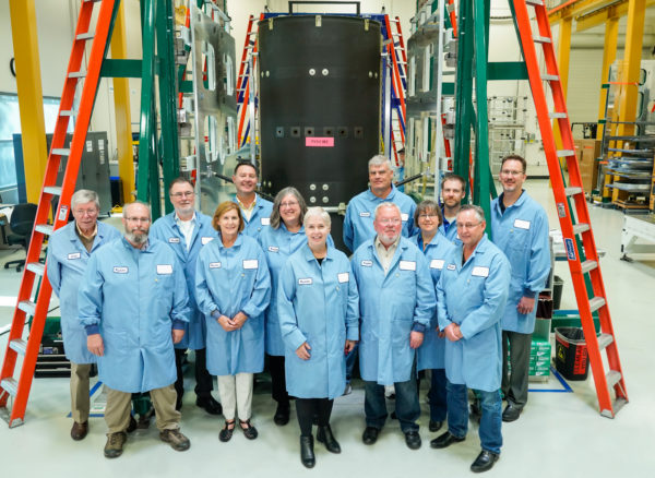 A group of people in light blue protective coats standing in front of a large mechanical structure.