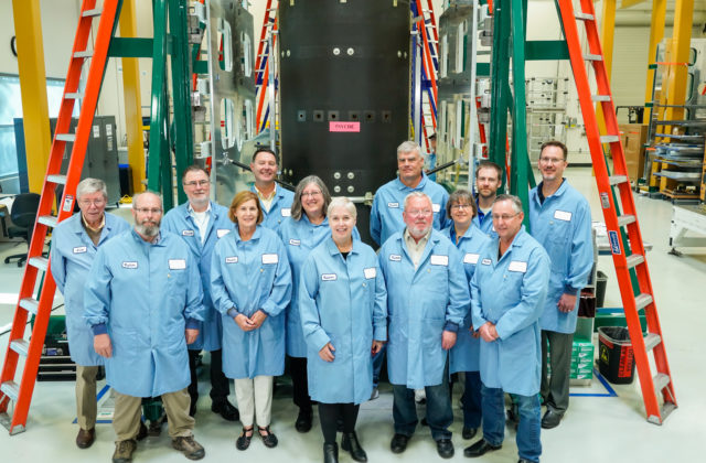 A group of people in light blue protective coats standing in front of a large mechanical structure.