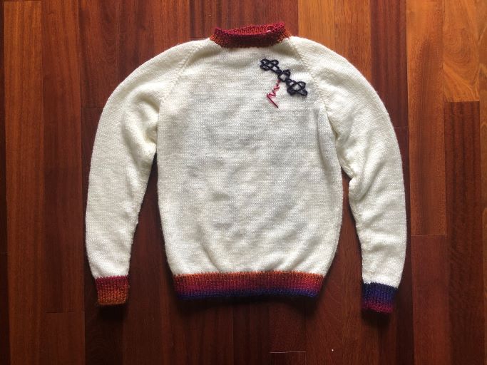 On the front, left side of a white sweater, there's a purple outline of the Psyche spacecraft with a small pink signal wave. On the back of the sweater is a large number 16 with the word “PSYCHE” spelled in all caps, placed like the design of a sports jersey. The collar, sleeve cuffs, lower band, letters, and numbers of the sweater are in an ombre yarn of purple, pink, red, and orange.