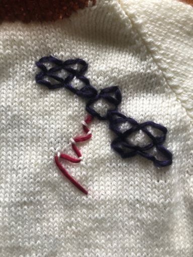 On the front, left side of a white sweater, there's a purple outline of the Psyche spacecraft with a small pink signal wave. On the back of the sweater is a large number 16 with the word “PSYCHE” spelled in all caps, placed like the design of a sports jersey. The collar, sleeve cuffs, lower band, letters, and numbers of the sweater are in an ombre yarn of purple, pink, red, and orange.