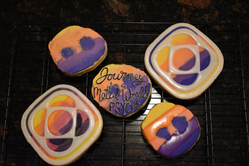 An asteroid-shaped cookie of the Psyche mission badge with black lettering that says, "Journey to a Metal World PSYCHE," is centered in the foreground. Mission badge and asteroid cookies of the same yellow, orange, pink, and purple gradient are in the background.