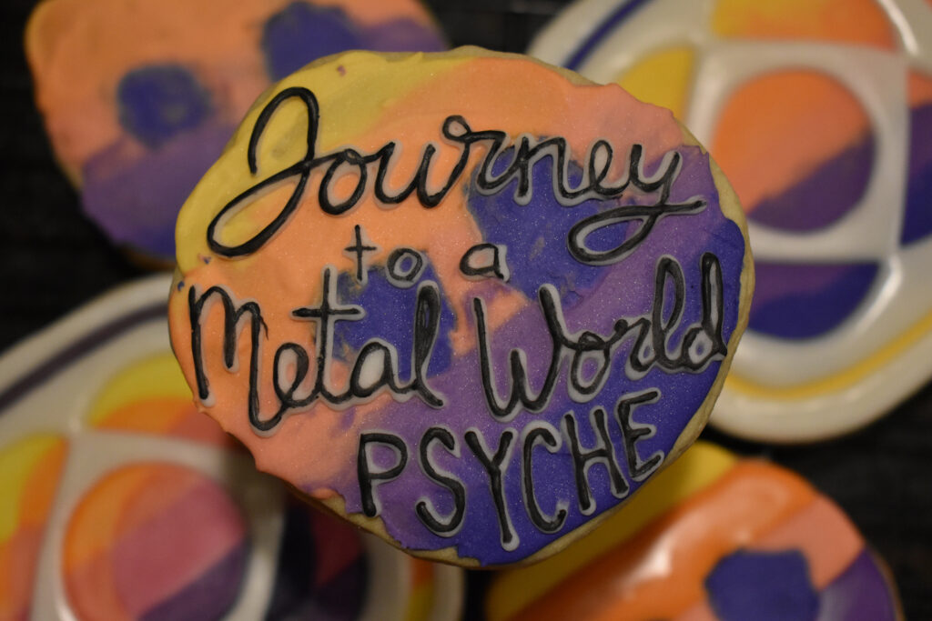 An asteroid-shaped cookie of the Psyche mission badge with black lettering that says, "Journey to a Metal World PSYCHE," is centered in the foreground. Mission badge and asteroid cookies of the same yellow, orange, pink, and purple gradient are in the background.