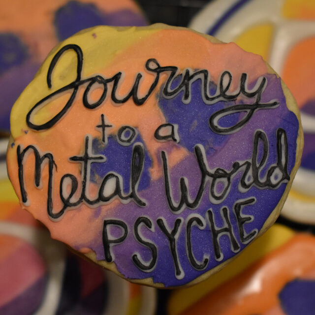 An asteroid-shaped cookie of the Psyche mission badge with black lettering that says, "Journey to a Metal World PSYCHE," is centered in the foreground. Mission badge and asteroid cookies of the same yellow, orange, pink, and purple gradient are in the background.