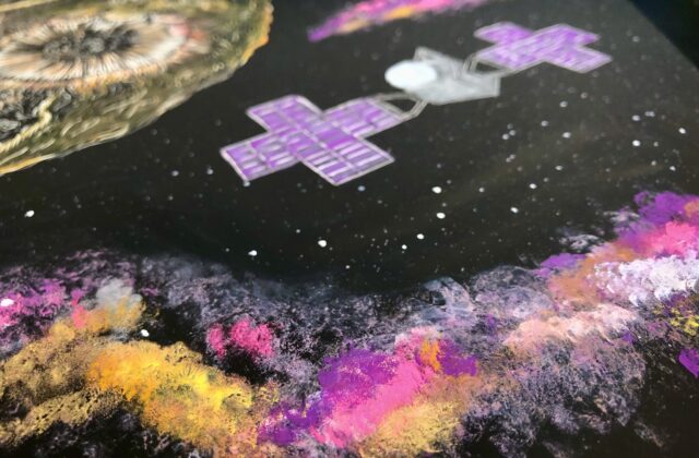 A 9 x 12 scratchboard embed with the drawing of Psyche and its satellite. To highlight the metallic texture and the colors of the Psyche mission, Psyche is contoured in a diluted gouache paint, and a more opaque form of the gouache paint in the mission's colors depict the satellite and the galaxy clouds.
