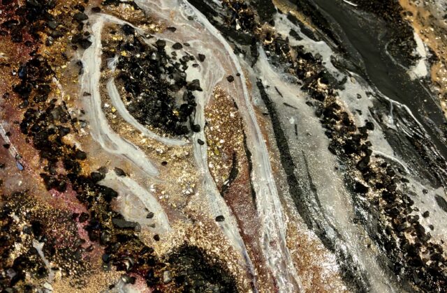 This abstract piece has gold, orange, and red streaks of paint on the bottom left corner with black crystals sprinkled over it. The paint is blended with black and white streaks. The black crystal textures and gold and silver glitter are present in most of the piece. The overall piece has a marble-like finish and appearance.