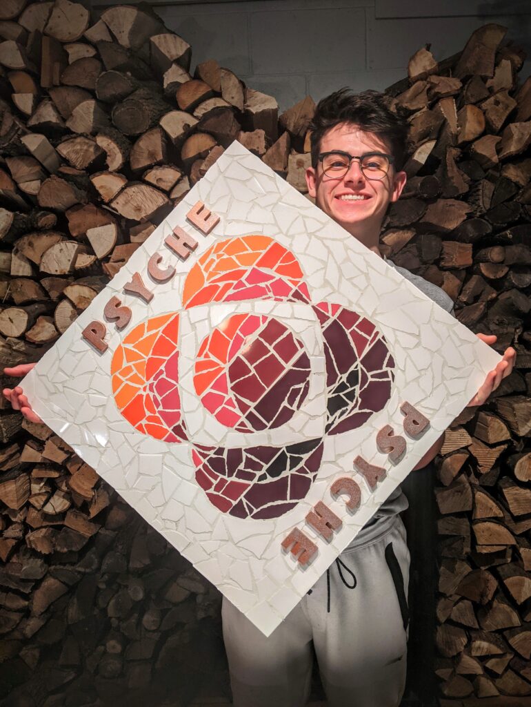 White, orange, pink, and purple ceramic tiles arranged in the shape of the Psyche logo with white grout finish and silver spray-painted wooden "Psyche" letters.