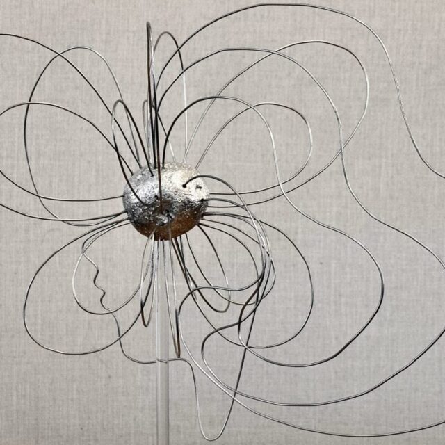 A shiny metal sculpture about 20 inches tall, composed of a model asteroid with many steel wires protruding from the surface. The wires loop back around into the asteroid, forming curved shapes that resemble a woman’s face and hair. The sculpture is supported by a clear rod connected to a marble base, making it appear to be floating freely.