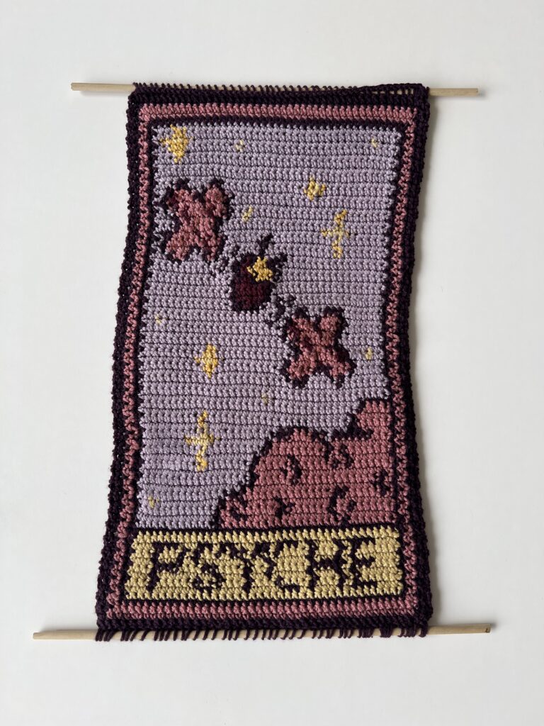 "Tapestry of Psyche" is a crochet tapestry blending medieval artistry with space exploration. Using yarns inspired by the NASA Psyche mission, it features the Psyche spacecraft, stars, and a whimsical cloud. A pixelated "Psyche" graphic adds a modern touch.